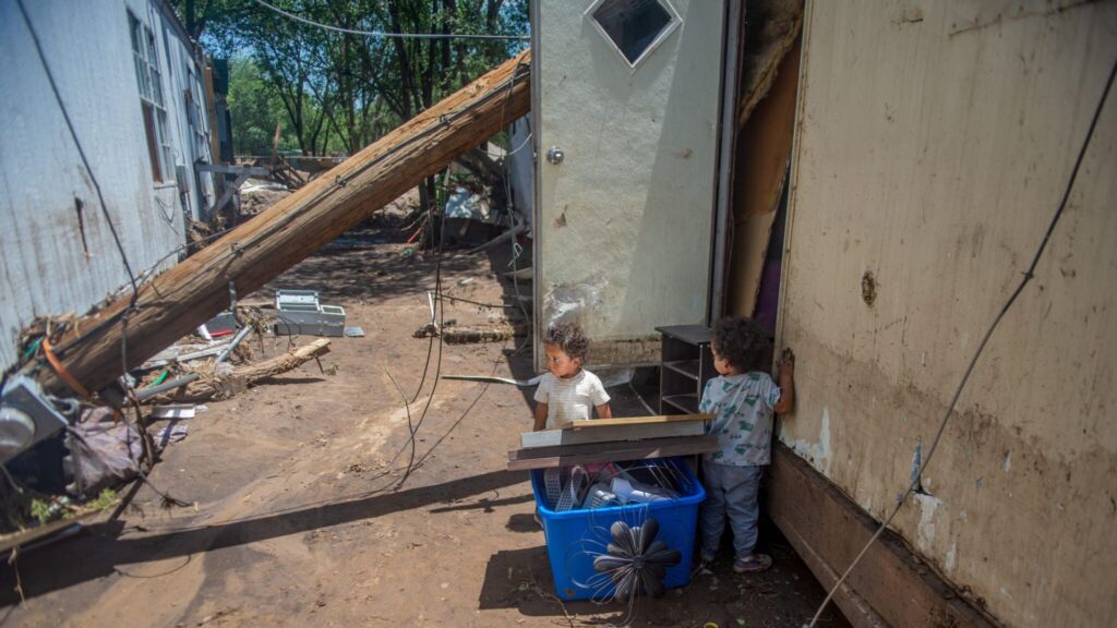 Two young children stand outside a damaged building with debris and a fallen utility pole nearby. The ground is muddy, and belongings are scattered in a plastic bin.