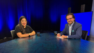 Two people sit at a blue table in a studio with blue lighting, facing the camera. One has a notepad and pen, the other is smiling. Two mugs are on the table.