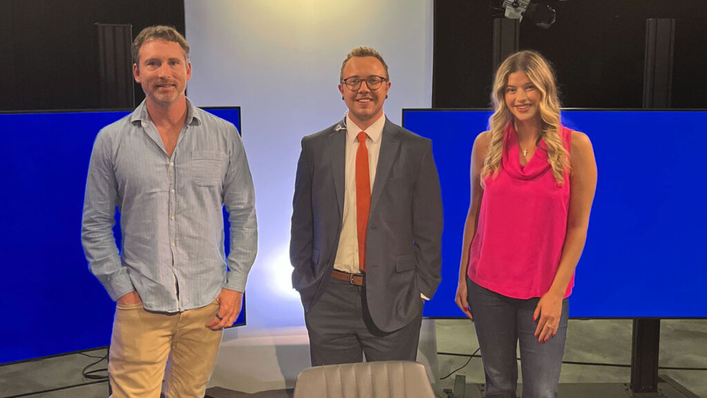 Three people stand indoors in front of large blue screens; one man in a light shirt and khakis, one man in a suit and tie, and one woman in a pink sleeveless top and jeans.