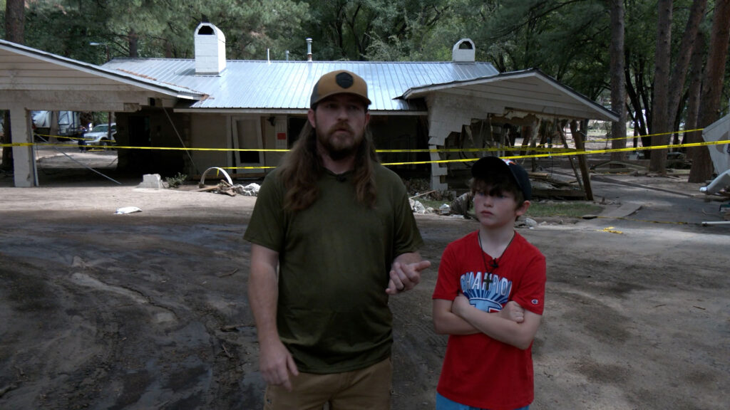 Two people stand in front of a damaged house with yellow caution tape across the area; debris and mud are visible on the ground.