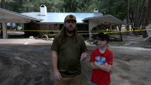 Two people stand in front of a damaged house with yellow caution tape across the area; debris and mud are visible on the ground.