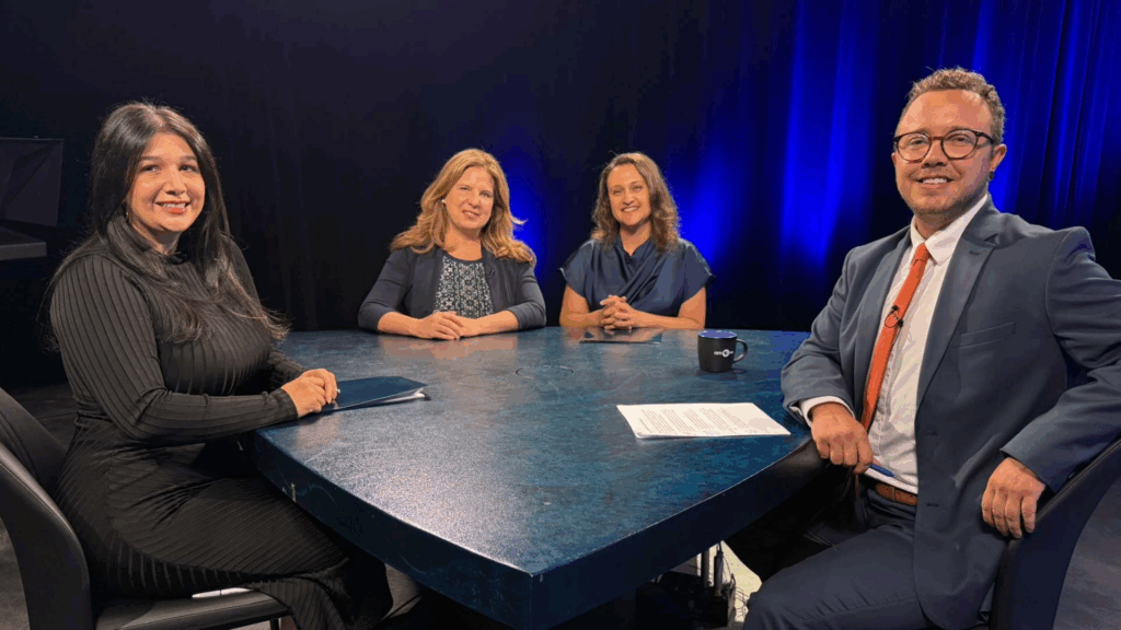 Four people, three women and one man, sit around a table in a studio setting with dark lighting and blue accents, appearing ready for a discussion or interview.