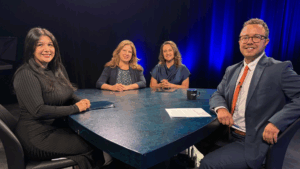 Four people, three women and one man, sit around a table in a studio setting with dark lighting and blue accents, appearing ready for a discussion or interview.