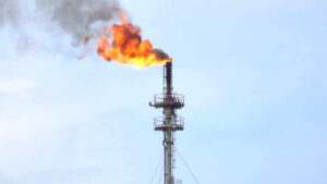 Tall industrial flare stack emitting a large orange flame and dark smoke against a pale blue sky.