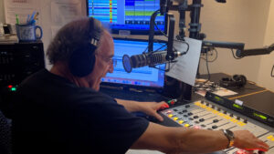 A man wearing headphones operates controls at a radio station mixing board, with multiple monitors and a microphone in front of him.