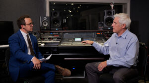 Two men sit and talk in a professional recording studio, with audio equipment and speakers visible in the background.
