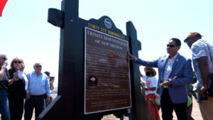 A group of people stand around a historical marker titled "Trinity Downwinders of New Mexico" as a man in a suit gestures toward the sign.
