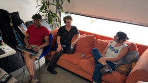 Three people are sitting in a casual office space; one works on a laptop while the other two converse on an orange couch.