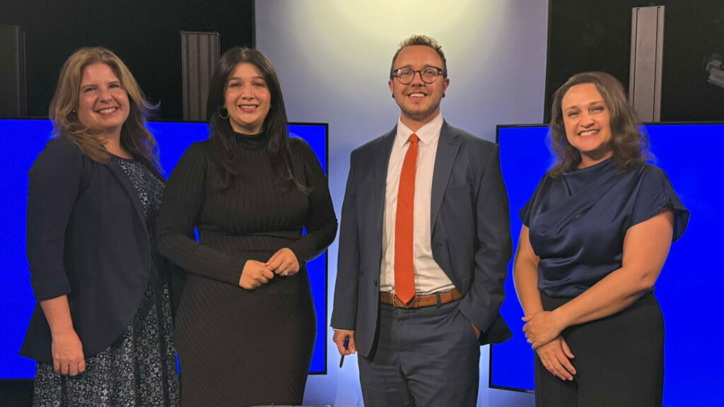 Four adults, three women and one man, pose and smile in front of blue screens in a studio setting. The man is wearing a suit and tie; the women are dressed in business or semi-formal attire.
