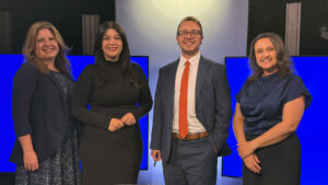 Four adults, three women and one man, pose and smile in front of blue screens in a studio setting. The man is wearing a suit and tie; the women are dressed in business or semi-formal attire.