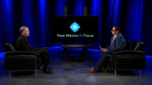 Two men sit in black chairs facing each other on a TV set with a screen displaying "New Mexico in Focus" and blue lighting in the background.