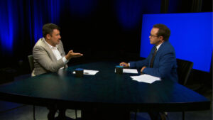 Two men in suits sit across from each other at a triangular table with papers and mugs, having a discussion in a studio with blue lighting and a blank blue screen in the background.
