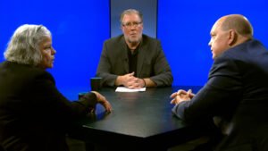 Three people in business attire sit around a black table having a serious discussion, with a blue background behind them.