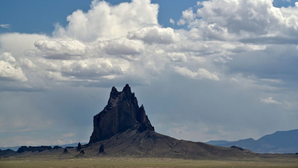 A jagged rock formation rises from a flat, arid landscape under a partly cloudy sky.