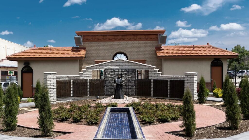 A courtyard garden with a statue in the center, surrounded by a low stone wall and a rectangular reflecting pool, in front of a building with a tiled roof.