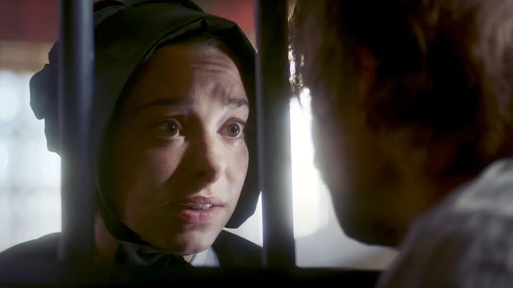 A woman in historical clothing and a bonnet looks closely at a man through metal bars, appearing concerned or emotional.