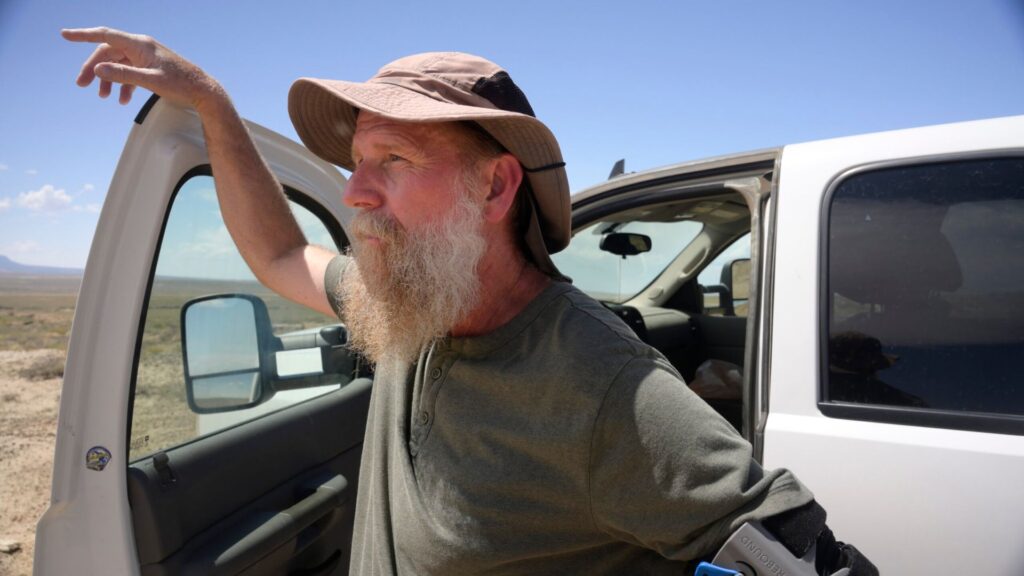 A man with a beard and sun hat leans on the open door of a white vehicle in a sunny, desert landscape.