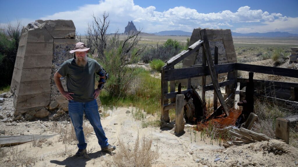 A man stands among abandoned, crumbling concrete and wooden structures in a dry, open landscape with distant rock formations under a partly cloudy sky.
