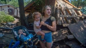 A woman holds a toddler and stands next to a baby in a stroller in front of a damaged house with debris scattered around.