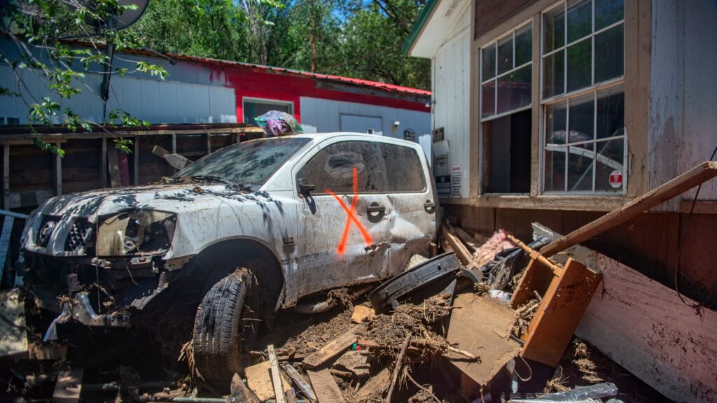 A damaged white pickup truck with a red X on the door sits among debris next to a house; the area shows signs of flooding and destruction.