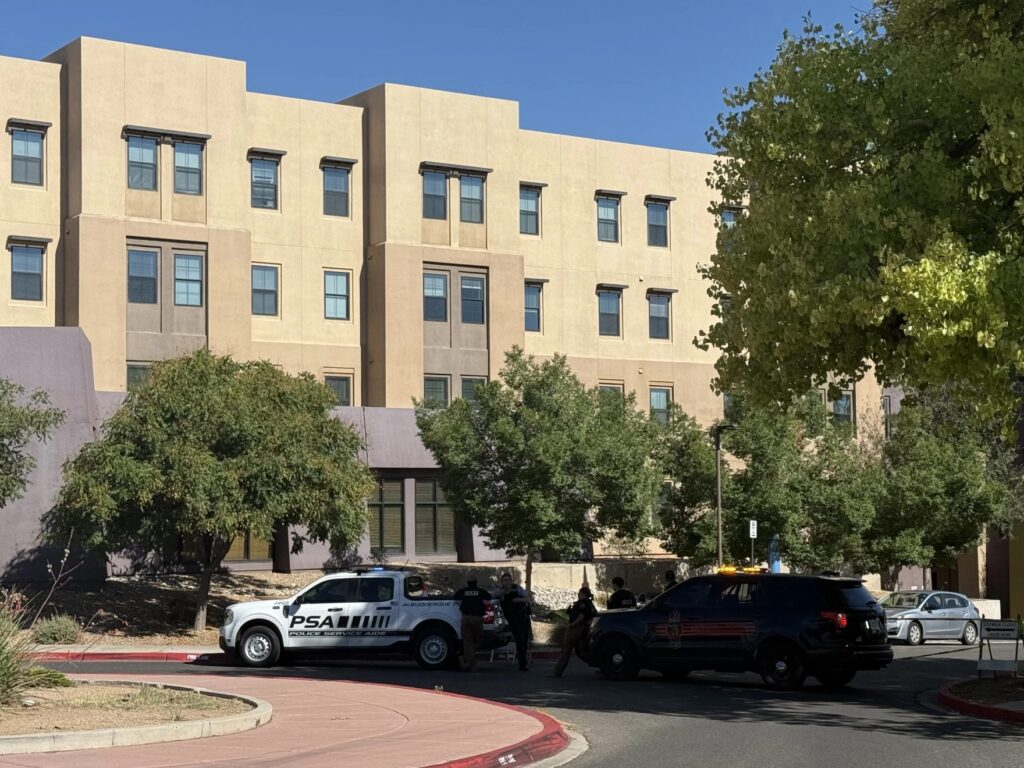 Two law enforcement vehicles and officers are stopped outside a multi-story building with trees and a clear sky in the background.