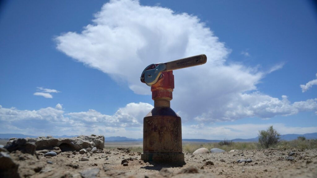 A rusty red irrigation valve stands in dry, sandy soil under a blue sky with scattered clouds in a desert landscape.