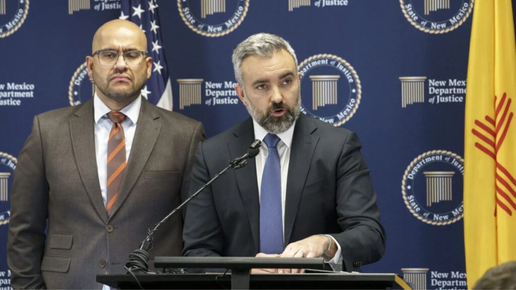 Two men in suits stand at a podium during a press conference, with New Mexico Department of Justice and state flag banners in the background.