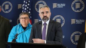 A man in a suit speaks at a podium with microphones, while another person stands beside him, against a backdrop with New Mexico Department of Justice logos and an American flag.