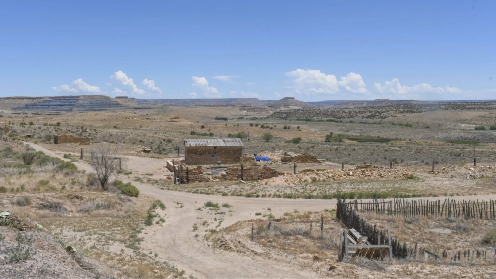 A solitary, weathered building sits surrounded by dry, sparse vegetation and dirt paths in a vast, arid desert landscape under a clear blue sky.