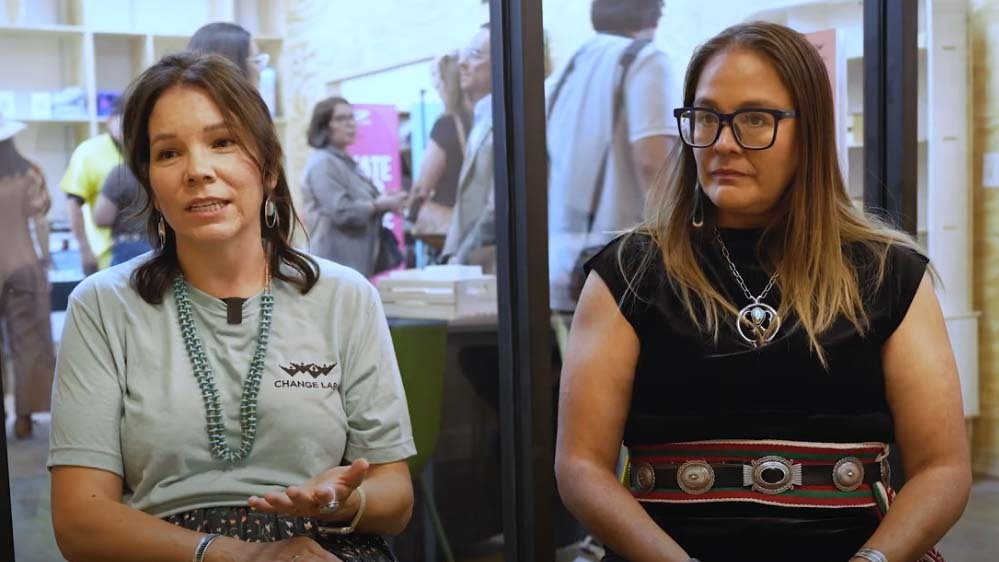 Two women sit indoors; one speaks with a hand gesture while the other listens. People and tables are visible in the background through a glass wall.