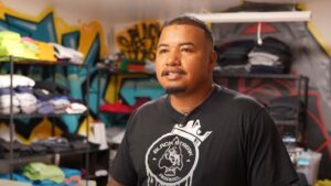 A man stands in a t-shirt shop with folded shirts on shelves behind him and colorful graffiti on the walls.