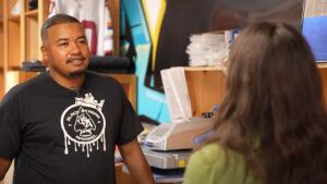 A man in a Black Street Modern t-shirt stands behind a counter, talking to a woman with long hair in a store or office setting.