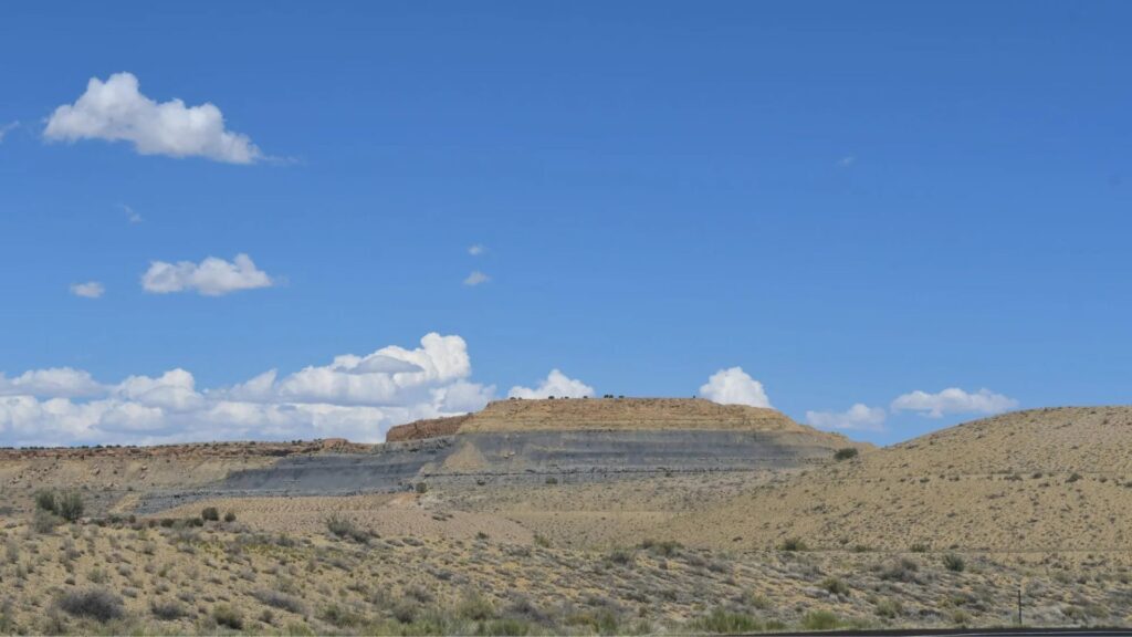 A flat-topped mesa rises above dry, scrub-filled land under a bright blue sky with scattered white clouds.