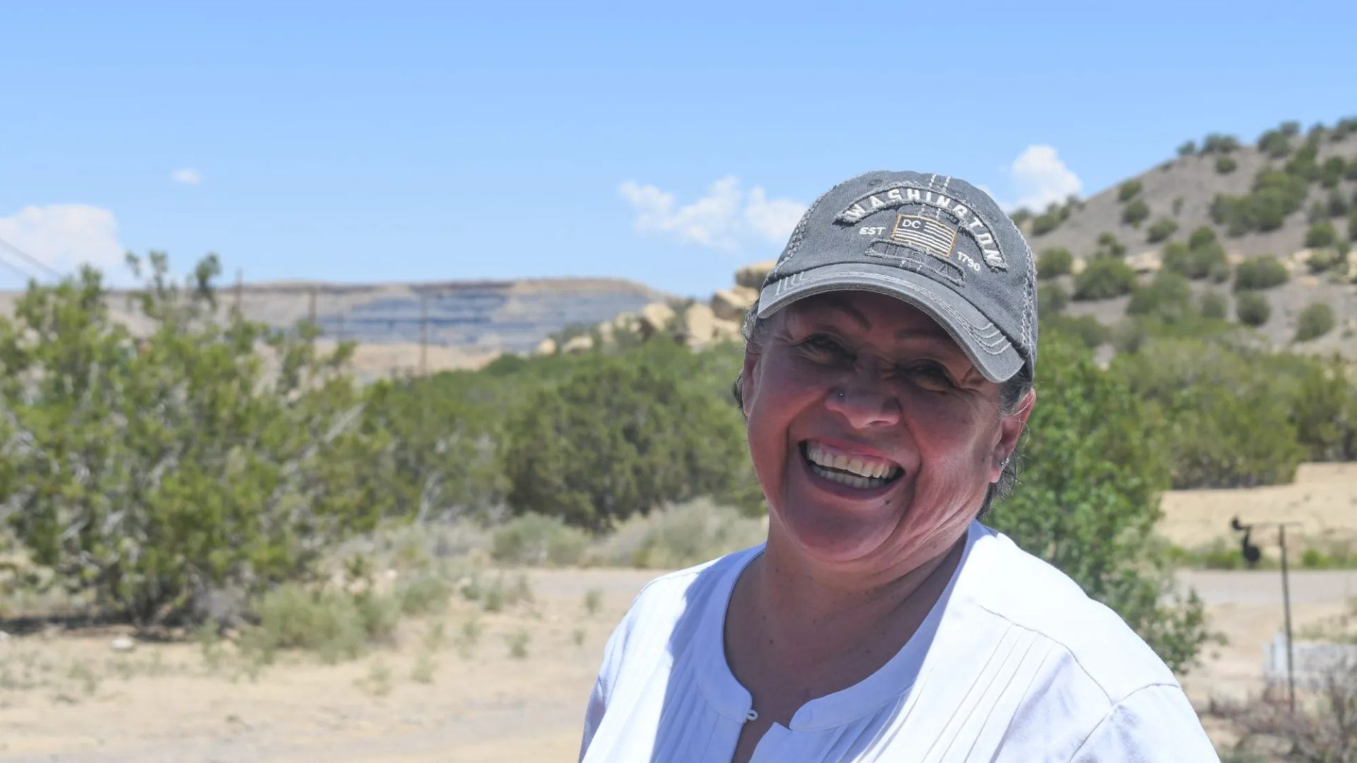 A woman wearing a gray cap and white shirt smiles outdoors in a desert landscape with bushes, hills, and a blue sky in the background.