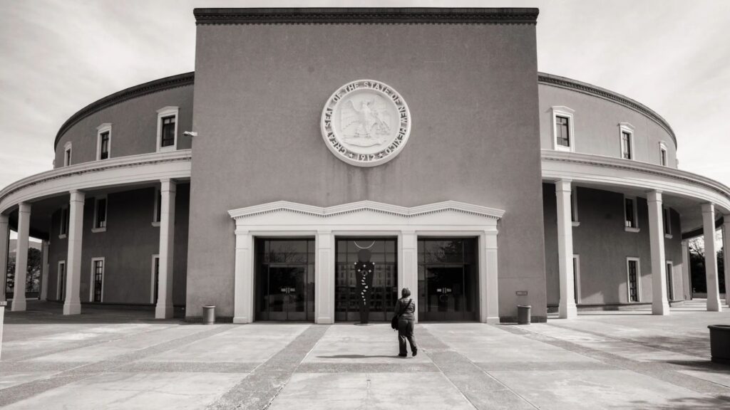 A person stands in front of the New Mexico State Capitol building, which features a circular design and a large seal above the entrance.