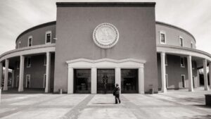 A person stands in front of the New Mexico State Capitol building, which features a circular design and a large seal above the entrance.