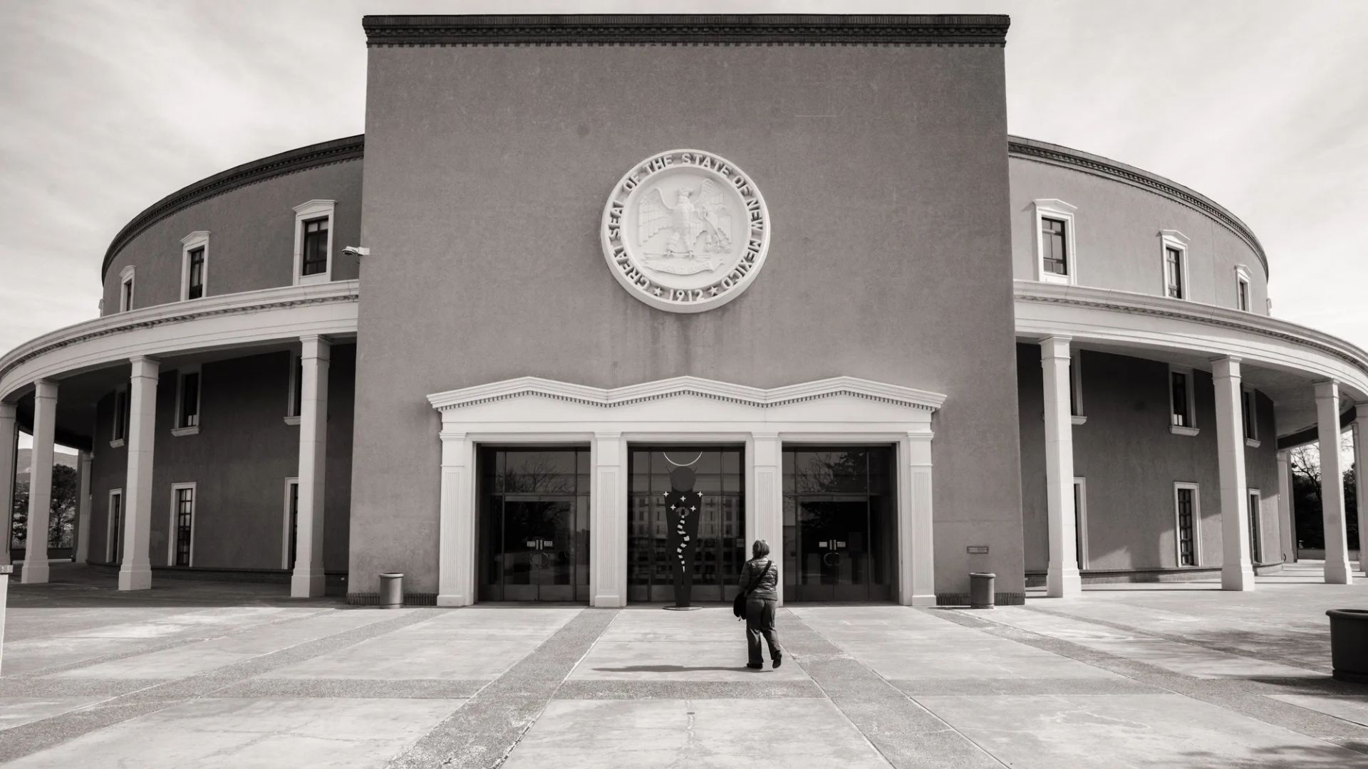 A person stands in front of the New Mexico State Capitol building, which features a circular design and a large seal above the entrance.