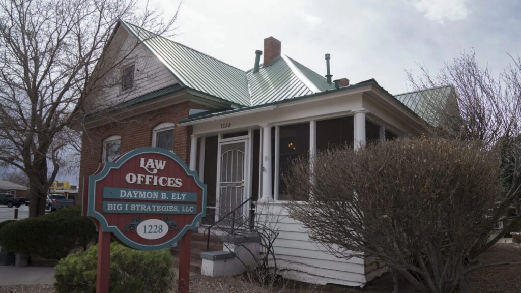 A small brick and white house with a green metal roof serves as a law office. A sign in front reads "Law Offices Daymon B. Ely, Big I Strategies, LLC, 1228." Bushes and trees surround the building.