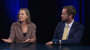 Two people sit at a table having a discussion; the woman on the left gestures with her hands while the man on the right listens attentively.