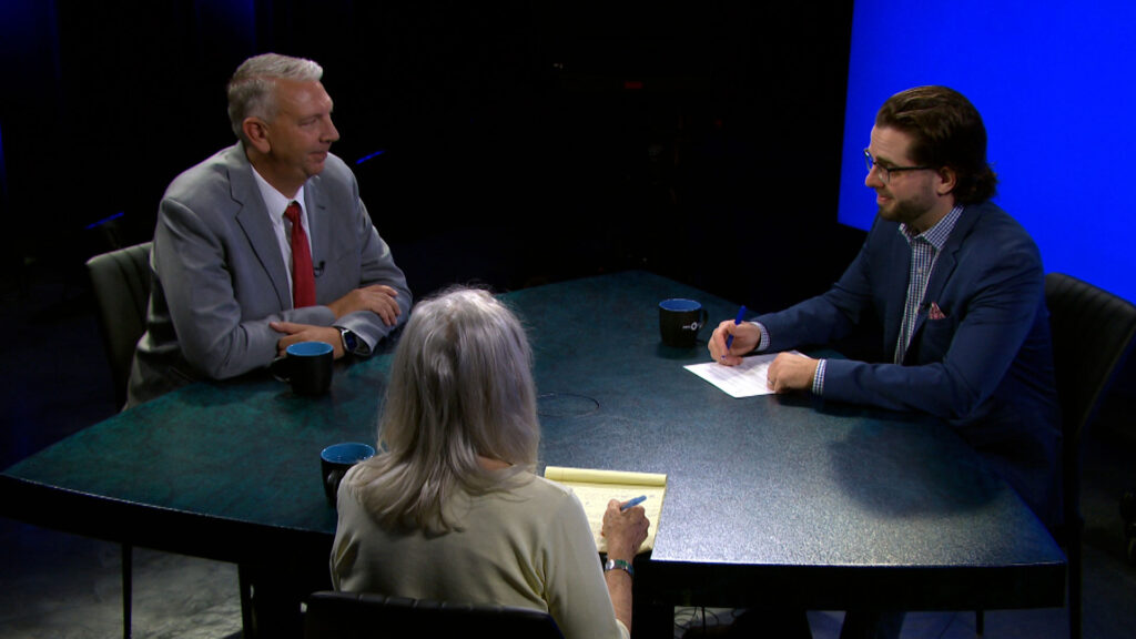 Three people sit around a table with mugs and notepads, engaged in a discussion in a studio with a blue background.