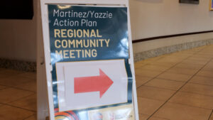 A sign reads "Martinez/Yazzie Action Plan Regional Community Meeting" with a red arrow pointing right. The sign is placed on a tiled floor indoors.