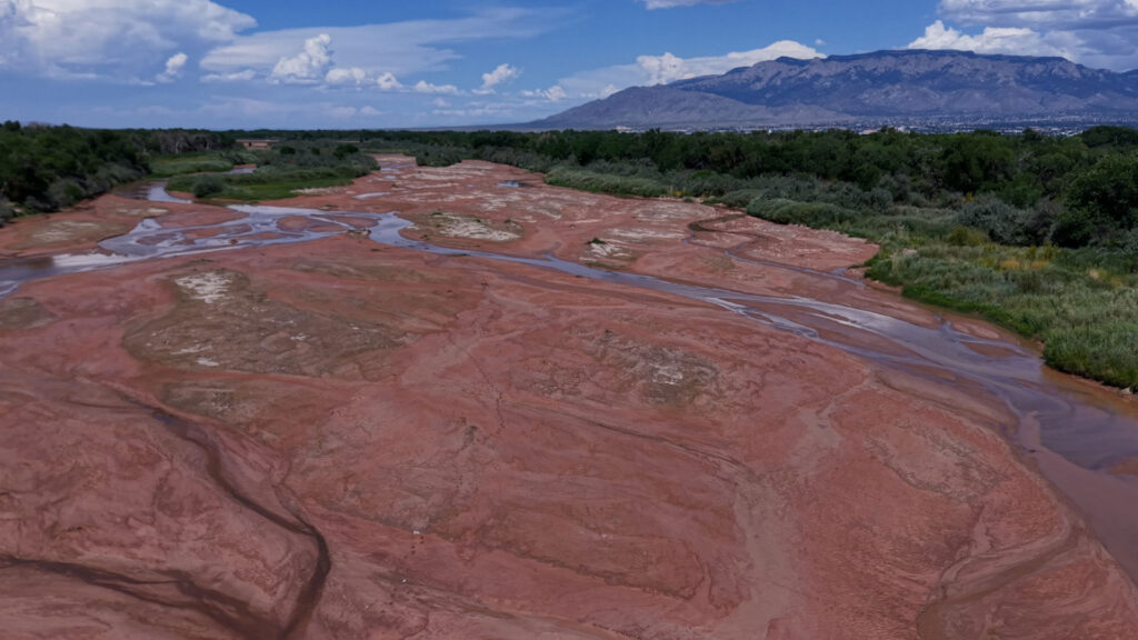 A mostly dry riverbed with scattered shallow streams, surrounded by green vegetation, under a partly cloudy sky with mountains in the background.