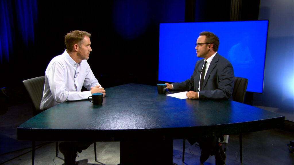 Two men sit across from each other at a triangular table, engaged in conversation, with coffee mugs and a large blue screen in the background.