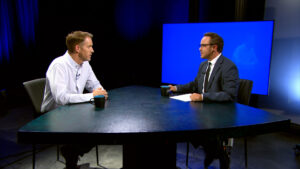 Two men sit across from each other at a triangular table, engaged in conversation, with coffee mugs and a large blue screen in the background.