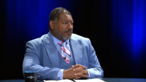 A man in a light blue suit and pink striped tie sits at a table with his hands clasped, next to a coffee mug, against a dark blue background.