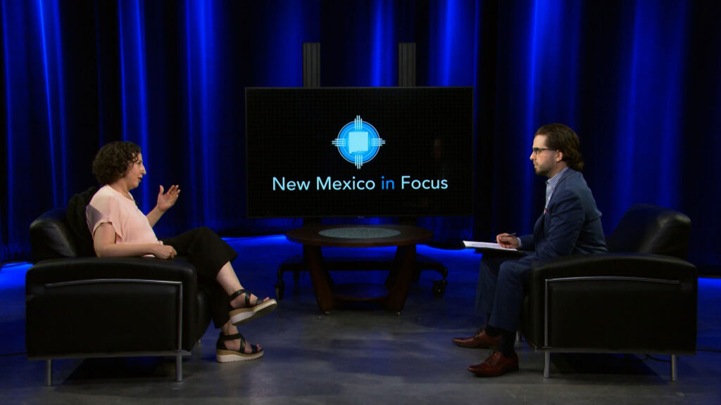 Two people sit across from each other in a studio with blue lighting, having a discussion. A screen between them displays the logo and text "New Mexico in Focus.