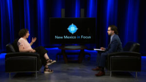 Two people sit across from each other in a studio with blue lighting, having a discussion. A screen between them displays the logo and text "New Mexico in Focus.