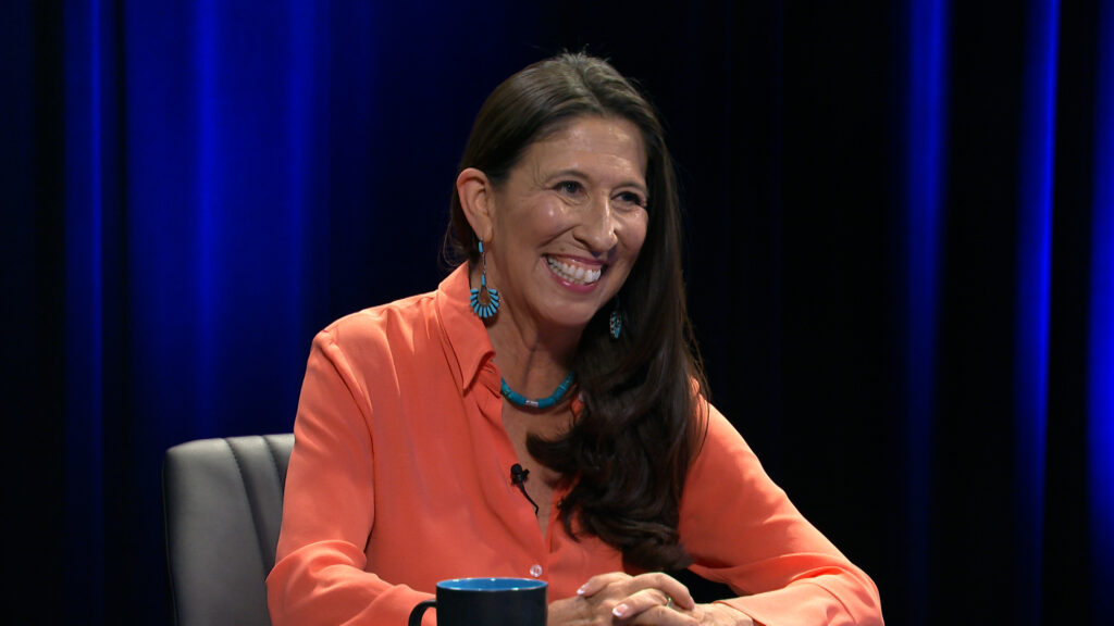 A woman with long dark hair, wearing an orange shirt and turquoise jewelry, sits smiling at a table in a studio with a dark blue background.