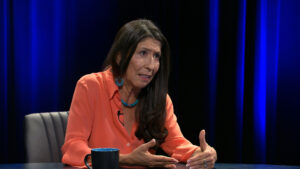 A woman with long dark hair, wearing an orange blouse and turquoise jewelry, sits at a table speaking with expressive hand gestures. A blue mug is in front of her.