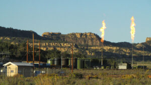 Two large gas flares burn above an industrial facility with storage tanks and buildings, set against a backdrop of rocky mesas and shrubland.